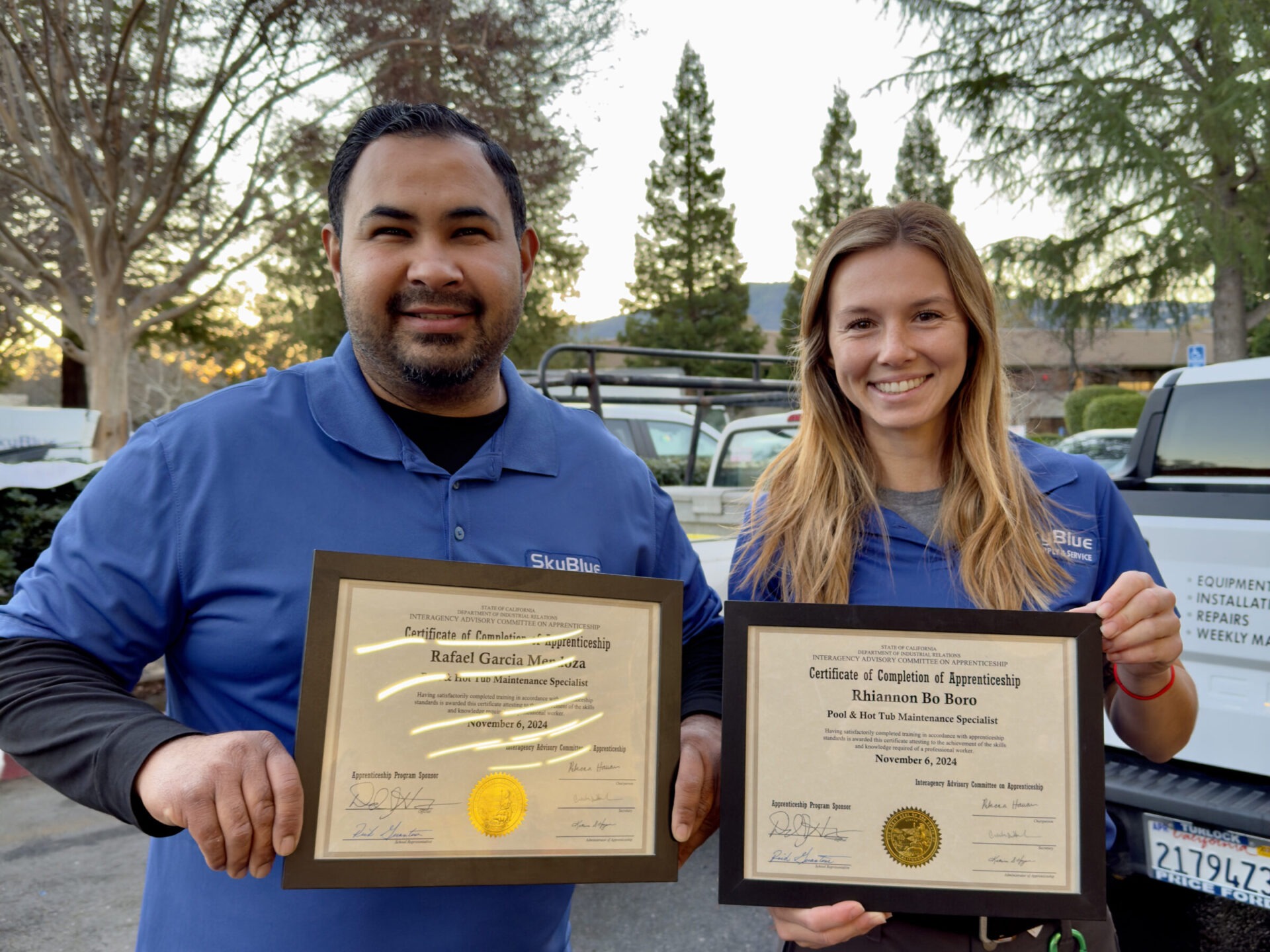 Two persons smiling, holding framed certificates outdoors. Background shows parked vehicles and tall trees. Both wear blue shirts, appearing proud of their accomplishments.