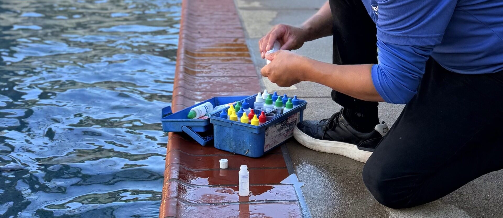 A person testing pool water with a kit, kneeling near the pool's edge. Various colored bottles are arranged in the blue container.