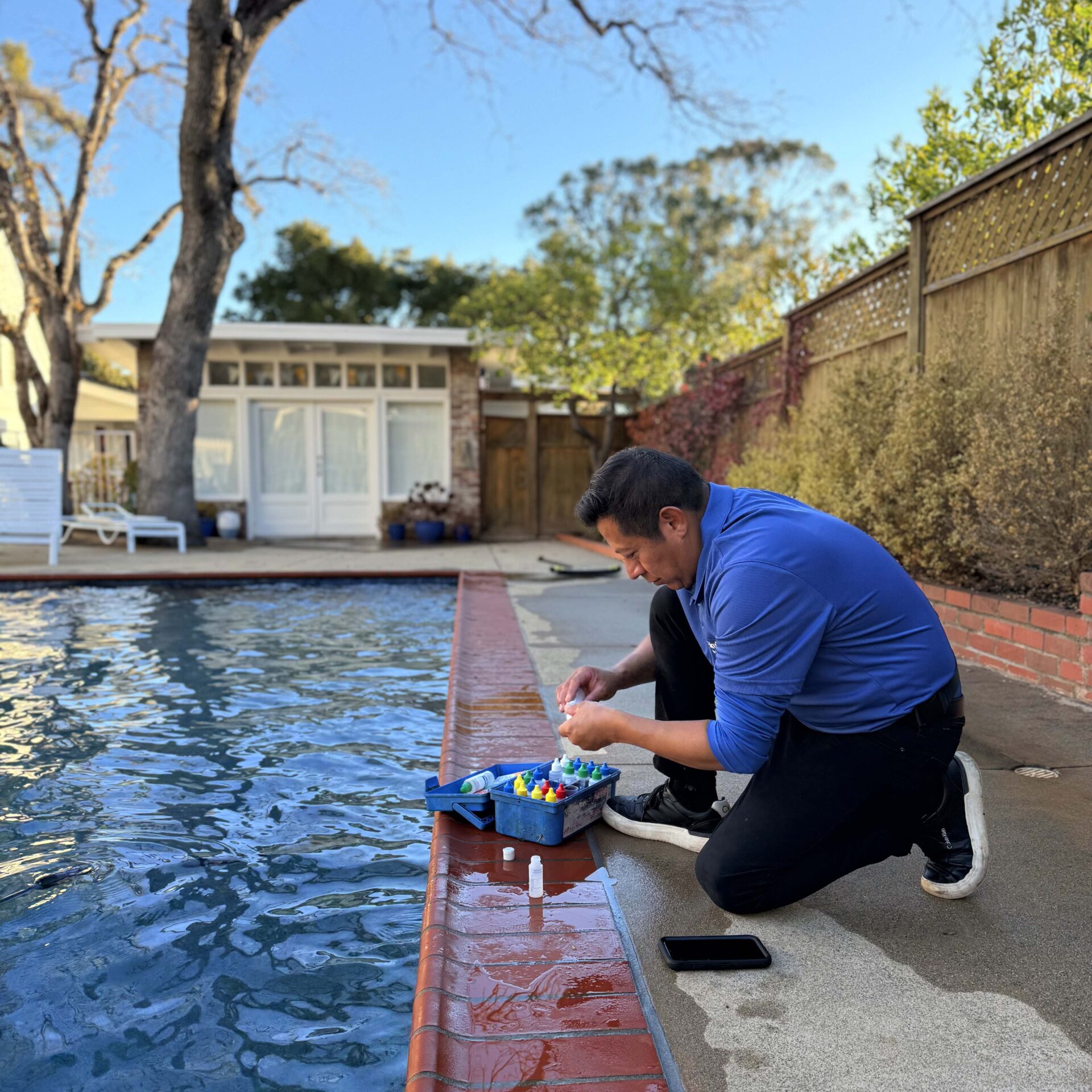 A person kneels by a swimming pool, testing the water with a kit beside a brick border, surrounded by trees and a fence.