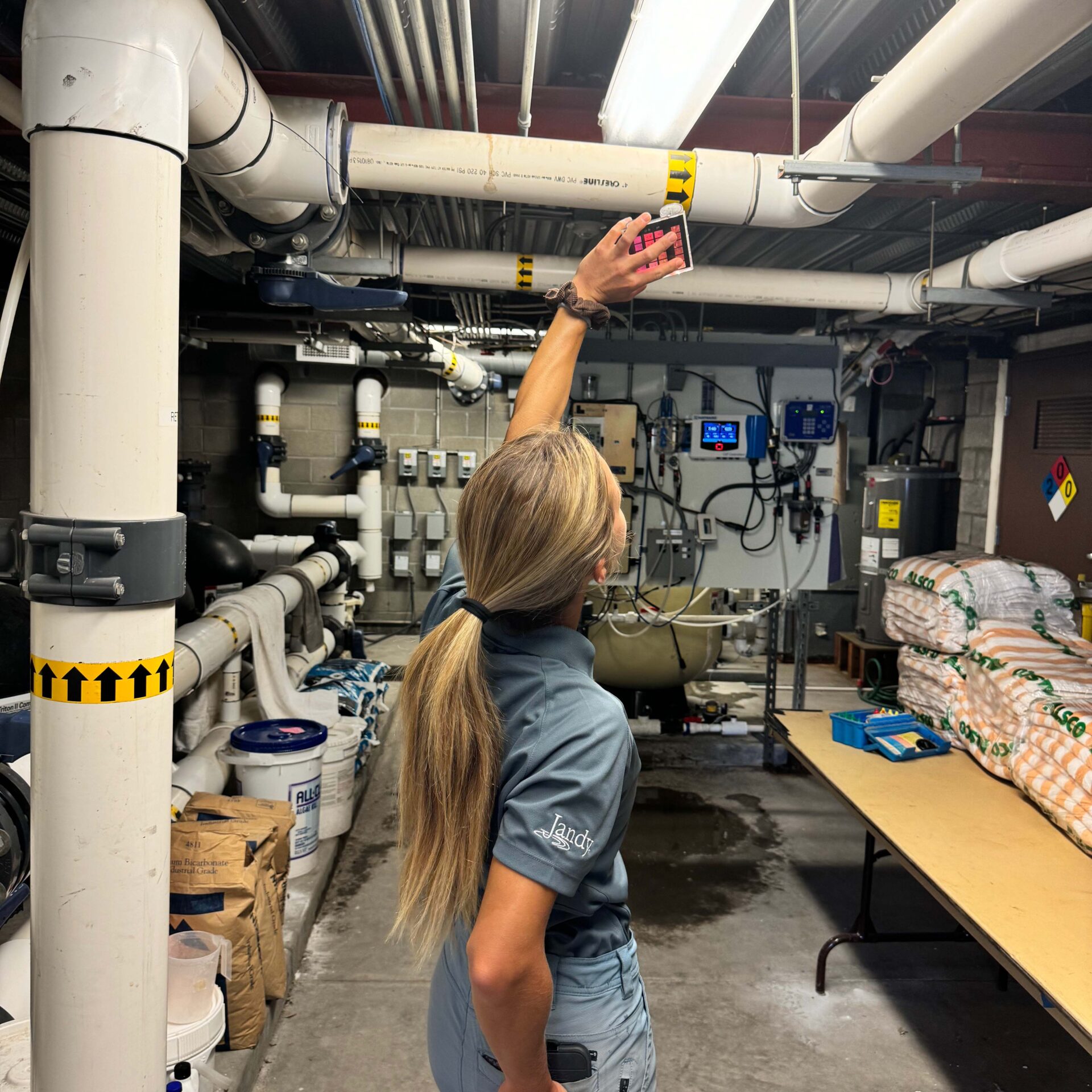 A person in a utility room points upward amidst pipes and equipment, conducting an inspection. Supplies are stacked nearby on a table.