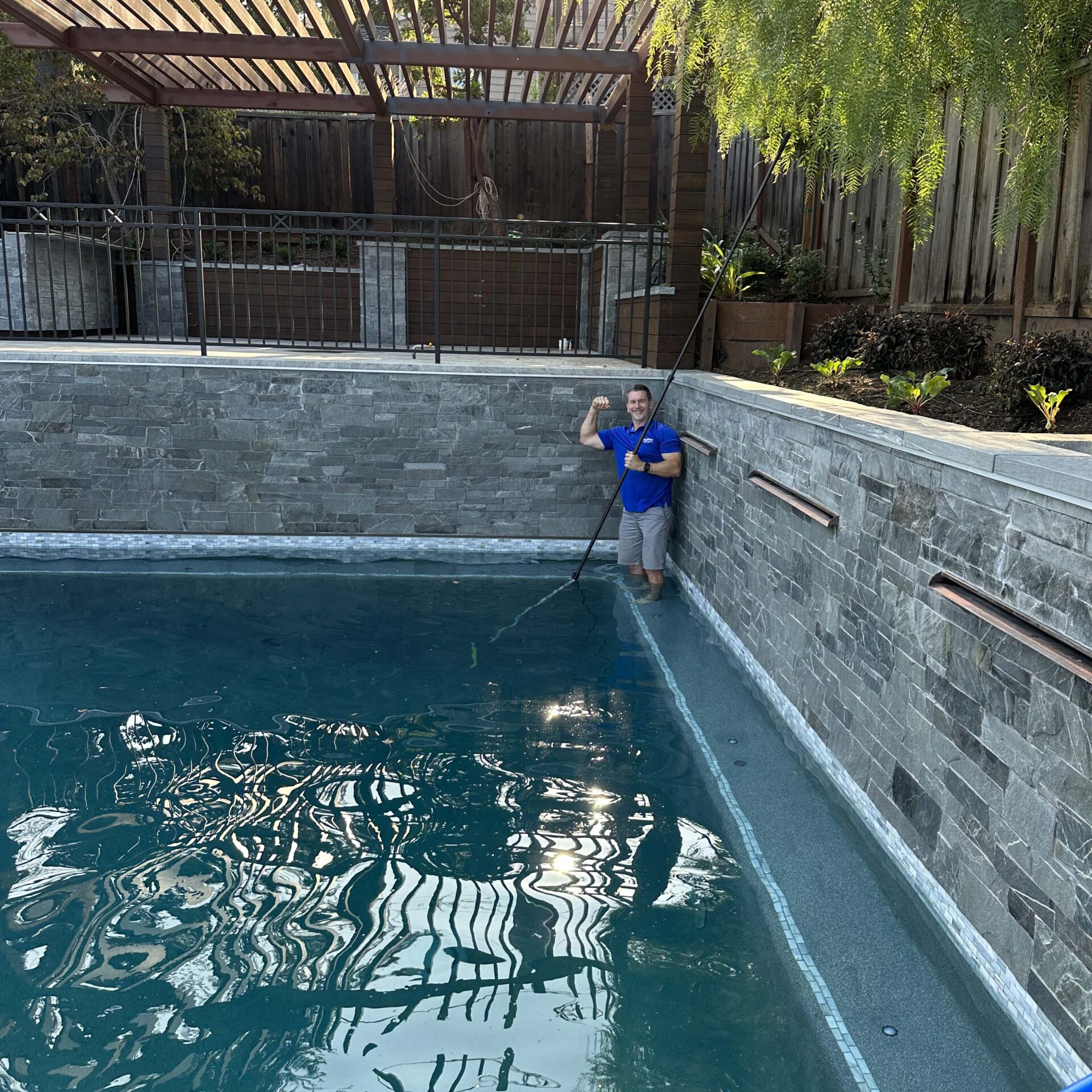 A person stands in an empty, stone-lined pool holding a cleaning tool. Wooden pergola and lush greenery are visible in the background.