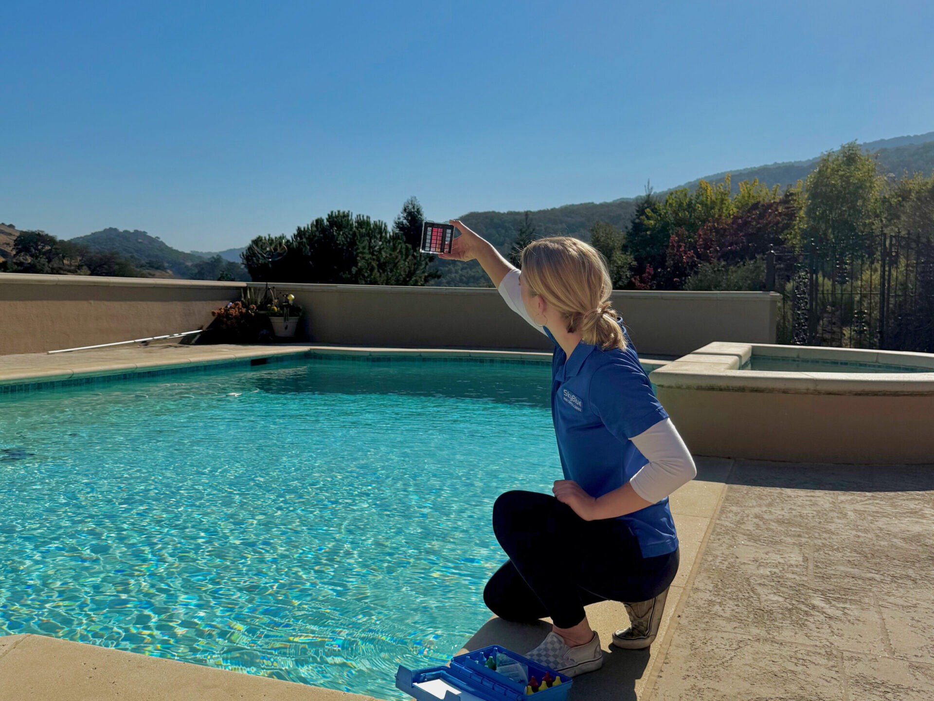 A person in blue attire tests swimming pool water with a kit, surrounded by mountains and greenery under a clear blue sky.