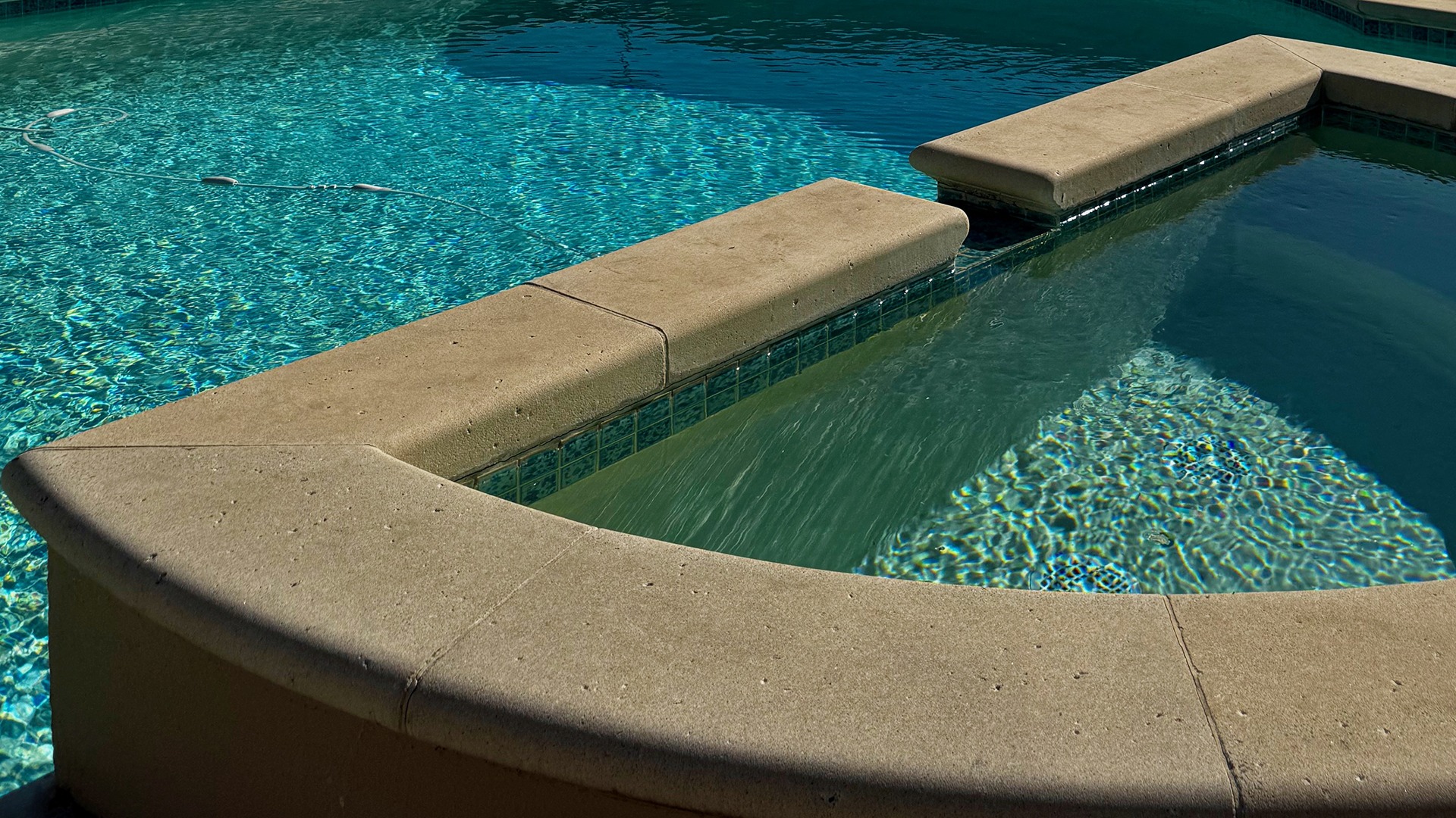 A close-up view of a sunny swimming pool with clean, inviting water and beige coping stones along the edge, creating contrasting textures.