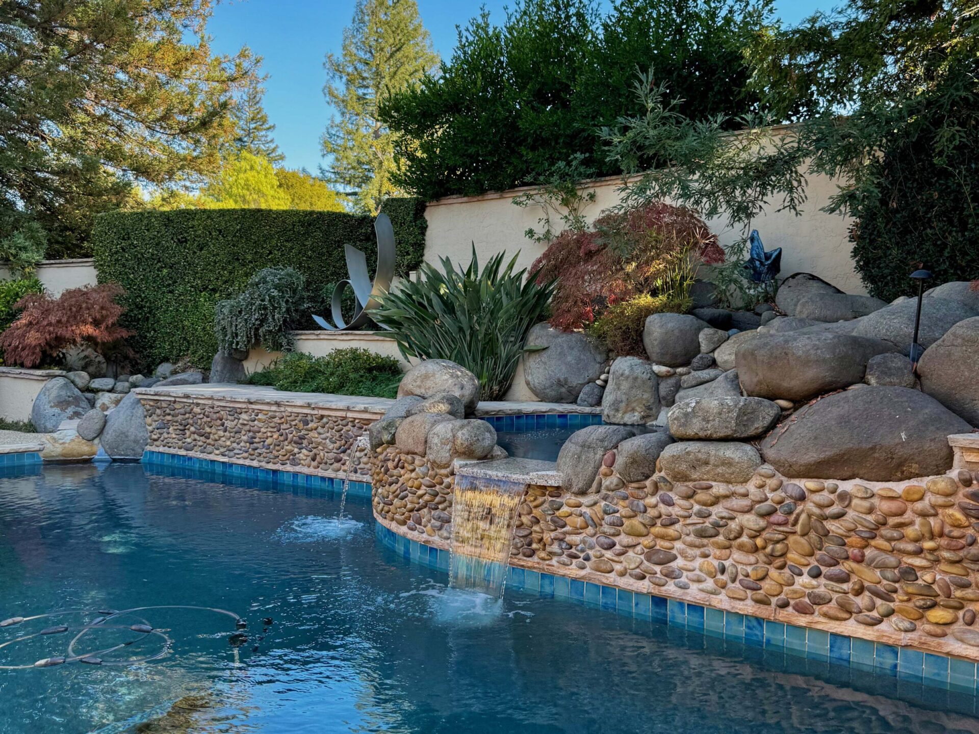 A serene garden pool with stone walls, cascading water, lush greenery, and carefully arranged rocks, creating a peaceful outdoor setting.