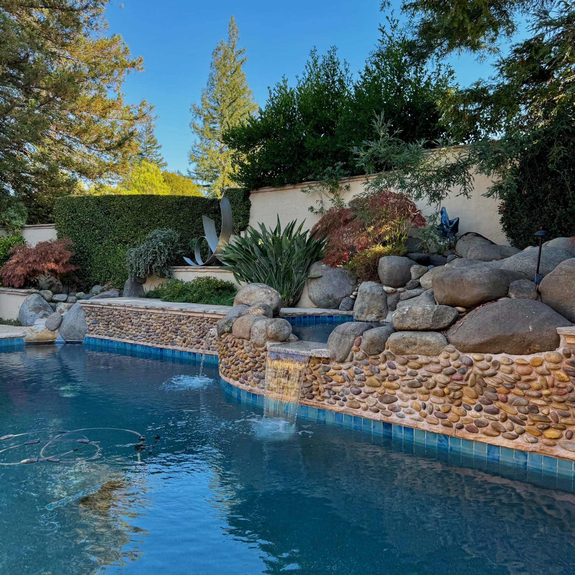 A backyard swimming pool with stone waterfall, surrounded by lush greenery and rocks, under a clear blue sky. No landmarks or people visible.