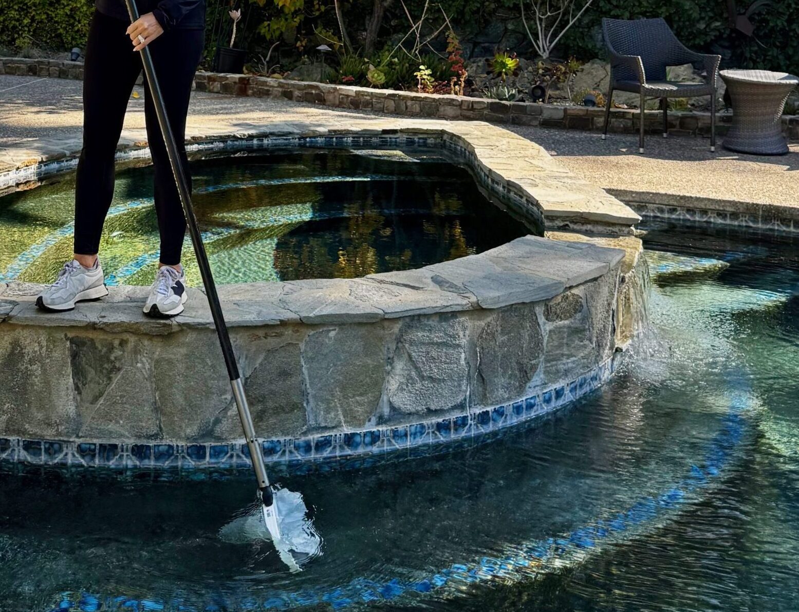 A person cleans a round stone-bordered pool with a net in a garden setting, surrounded by patio furniture and greenery.