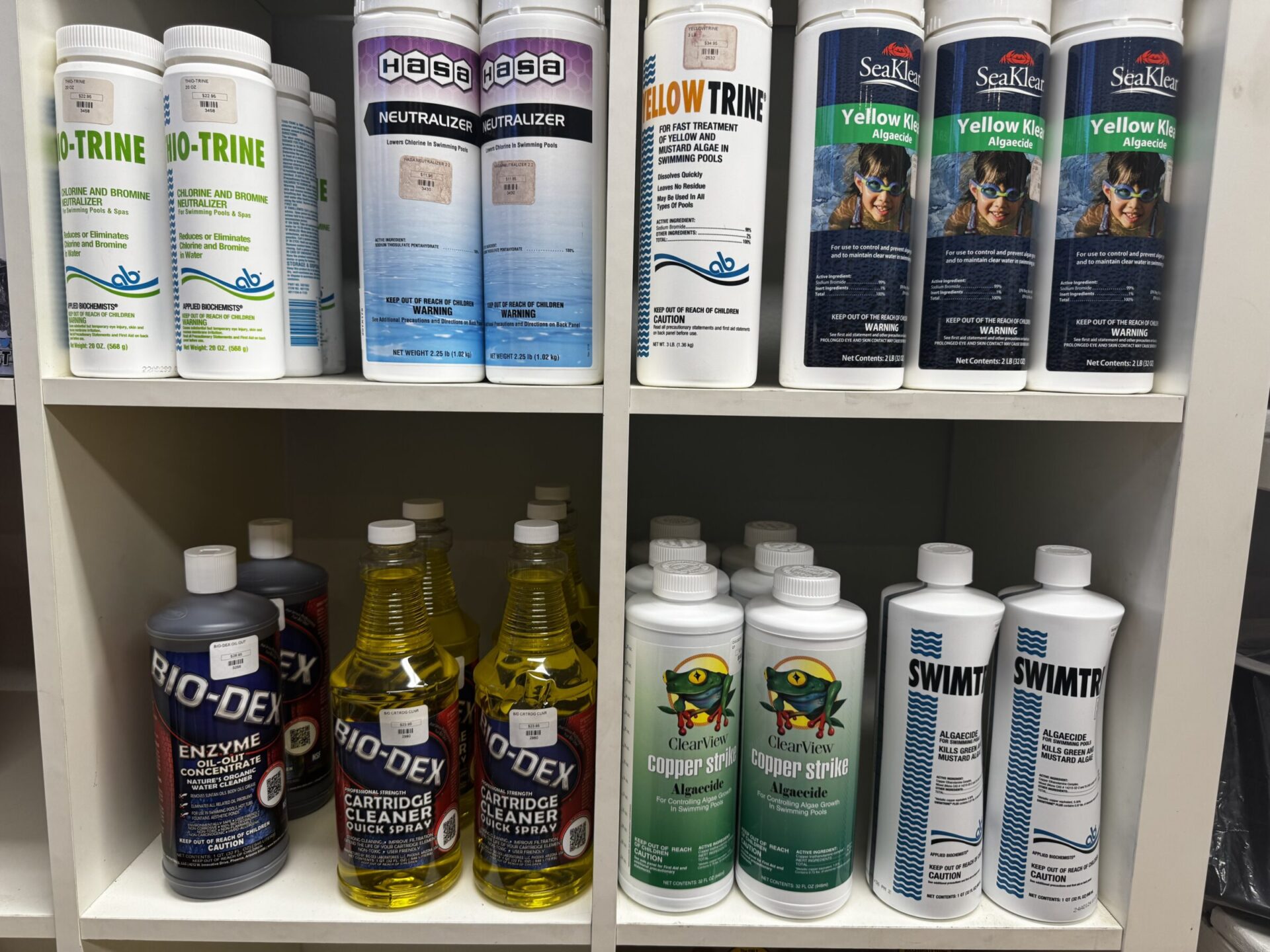 Shelves filled with various pool maintenance products, including enzymes, cleaners, and neutralizers, organized in rows. Brightly colored packaging and labels are visible.