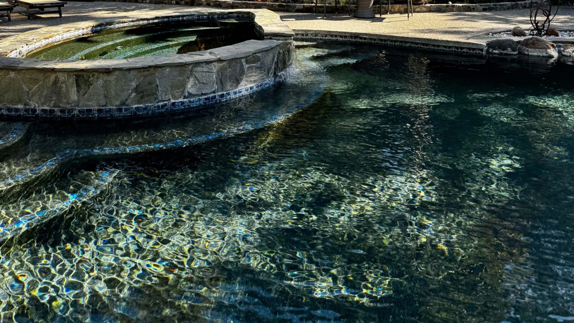 A crystal-clear pool with stone edges, surrounded by trees and sunlight reflections. No recognizable landmarks or historical buildings are visible.