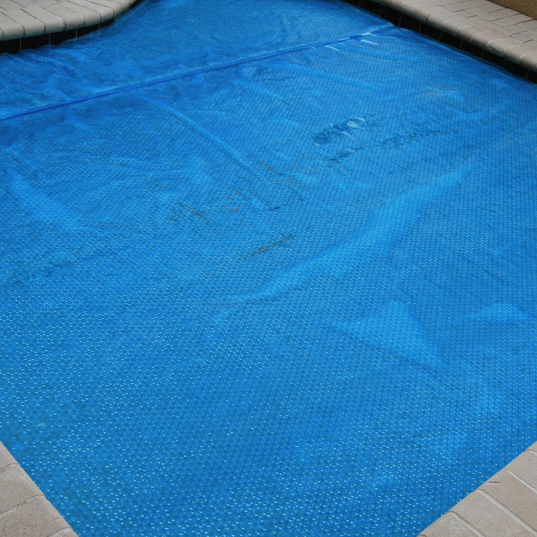 A covered swimming pool with a blue solar blanket. The surrounding area features light-colored paving, creating a clean and organized appearance.