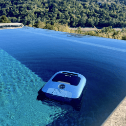 A robotic pool cleaner floats in a serene infinity pool overlooking a lush, green forest landscape under a clear blue sky.