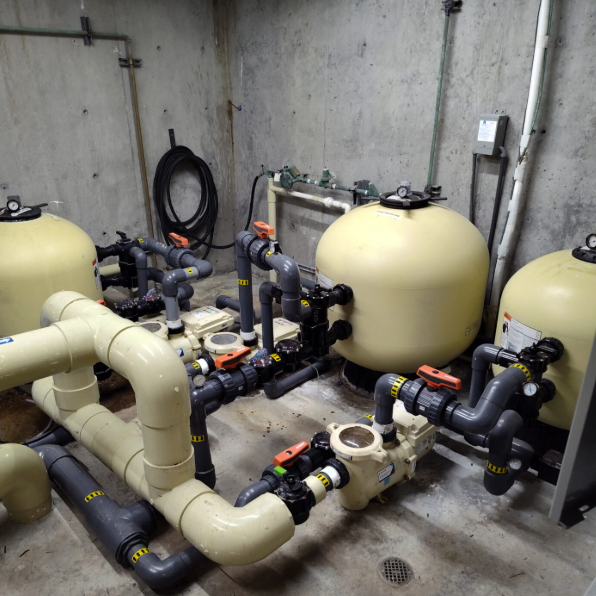 A basement room with large water filtration tanks and complex piping system against concrete walls. No people or recognizable landmarks are present.